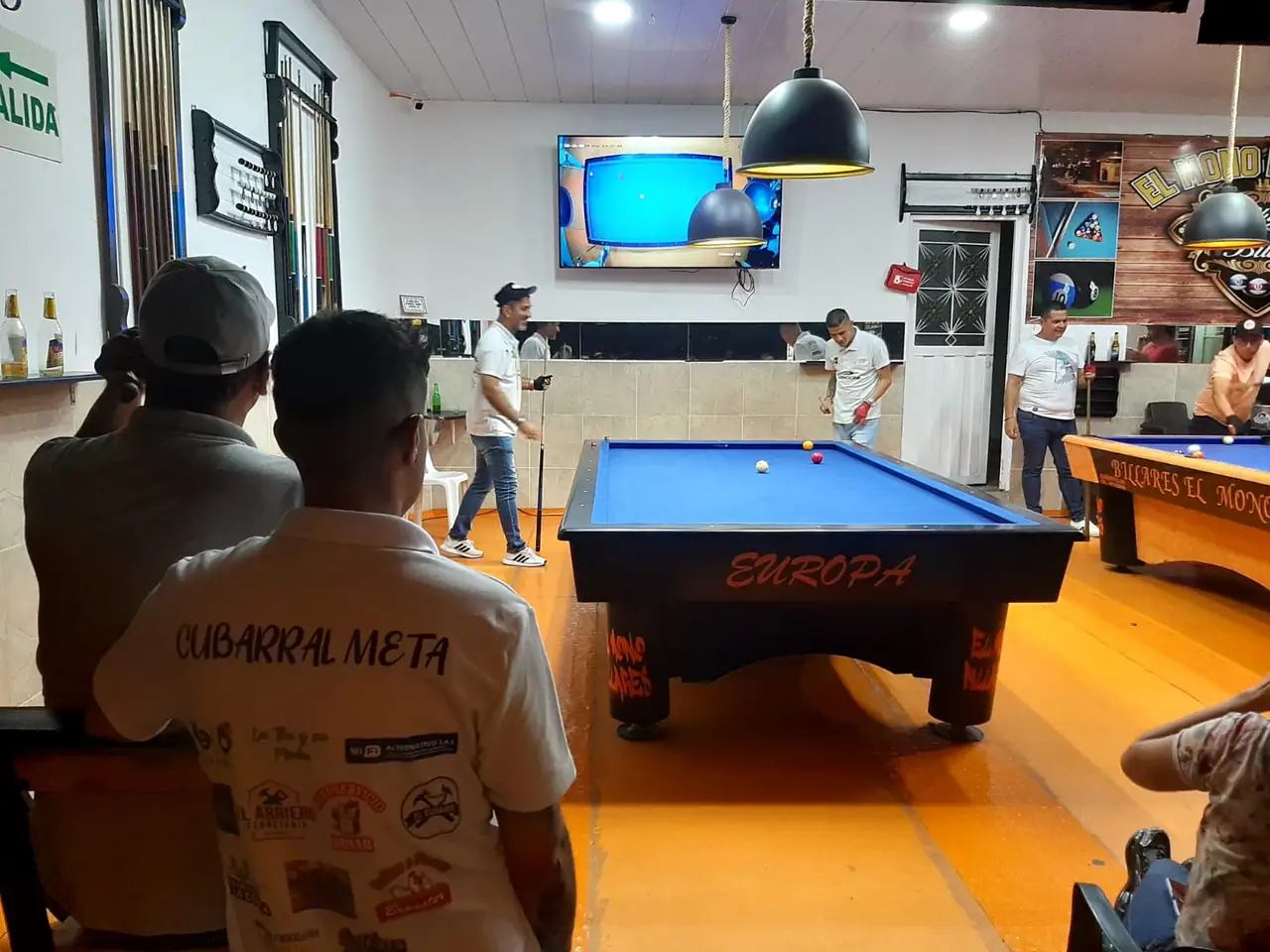 Público siguiendo una partida de billar; un asistente lleva una camiseta que dice Cubarral, Meta