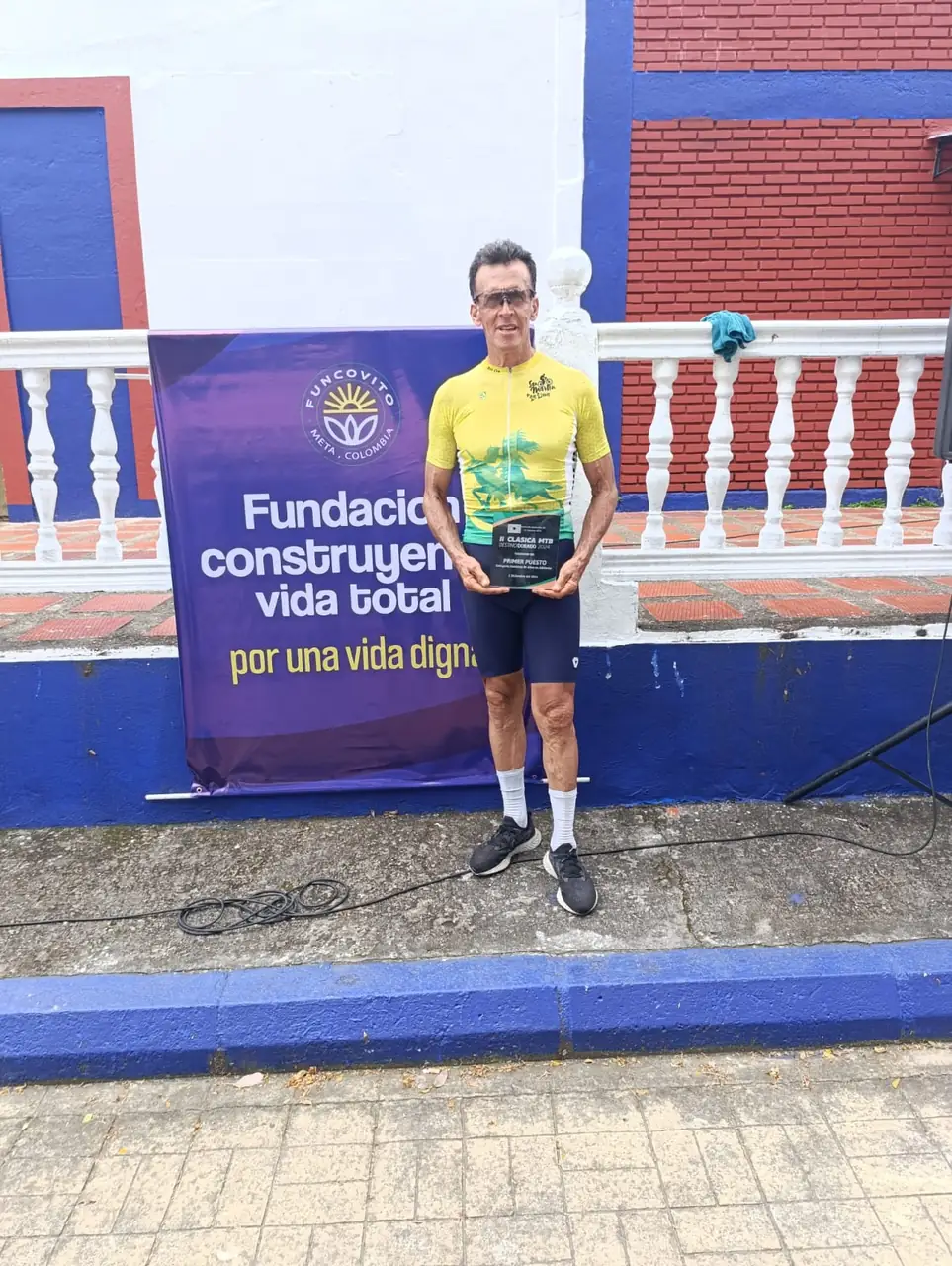 Ciclista con camiseta amarilla y su placa frente al pendón de la fundación